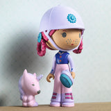 A doll with pink hair and a white helmet, holding a blue brush, stands beside a small purple unicorn. The doll wears a blue and pink outfit.