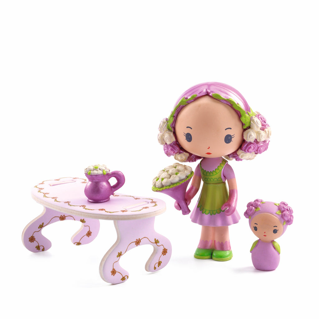 Toy figurines in pastel colors: one doll with pink hair and green shoes holding flowers, another smaller doll, and a pink table with a floral design.