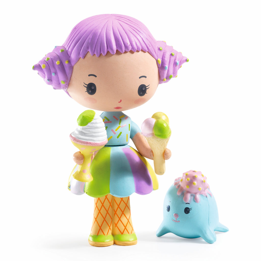 A cute doll with purple hair, holding an ice cream and dessert, wears a colorful dress and stands next to a smiling, blue seal-like creature.