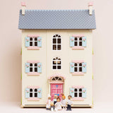 Seven wooden family figures standing in front of a light yellow dollhouse with pink doors and shutters.
