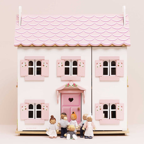 Seven wooden family figures gathered in front of a white dollhouse with pink shutters and a scalloped roof.
