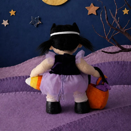 Back view of a plush doll in a purple dress, holding a pumpkin bag and candy corn against a starry night background.