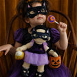 Young girl in a cat mask holding a matching plush doll and Halloween accessories in a wooden chair.