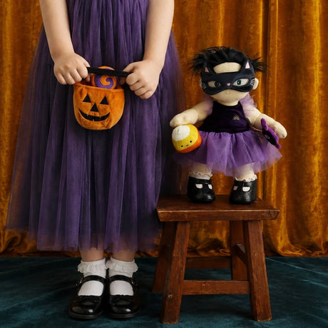 Plush doll on a wooden stool beside a child in a purple dress with a pumpkin bag.