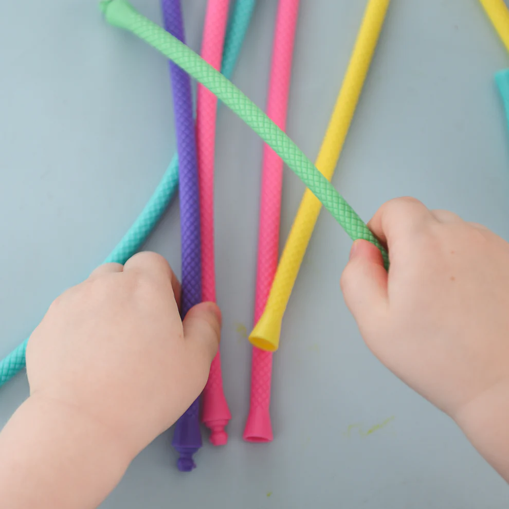 Child's hands eagerly grasp colorful, textured pop tubes in blue, purple, pink, and yellow on a light surface, conveying a playful, tactile experience.
