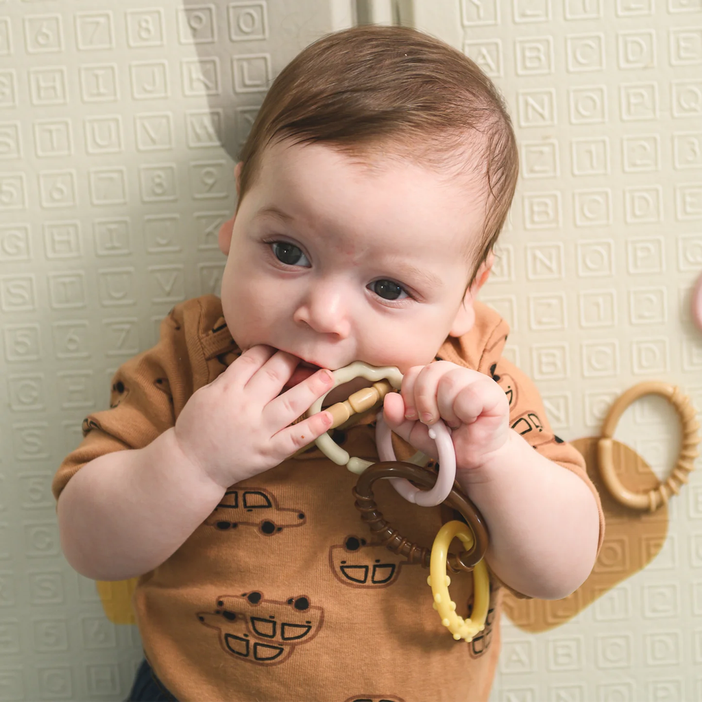 A baby in a brown shirt with car patterns chews on a colorful ring toy. The background features a textured, lettered play mat, conveying a playful mood.