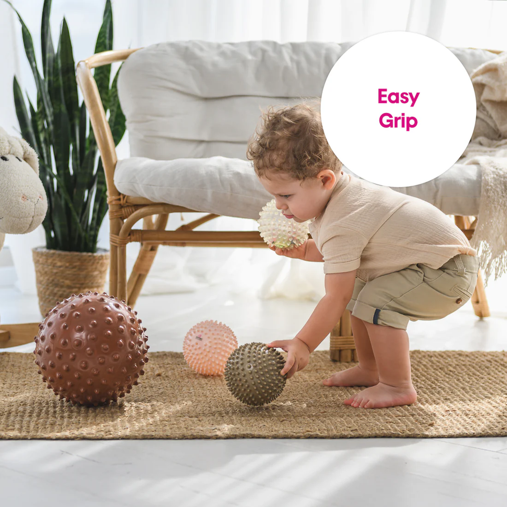 A toddler plays with four textured balls on a woven rug in a bright room. Behind them is a comfy rattan chair and green plant. Text reads "Easy Grip."