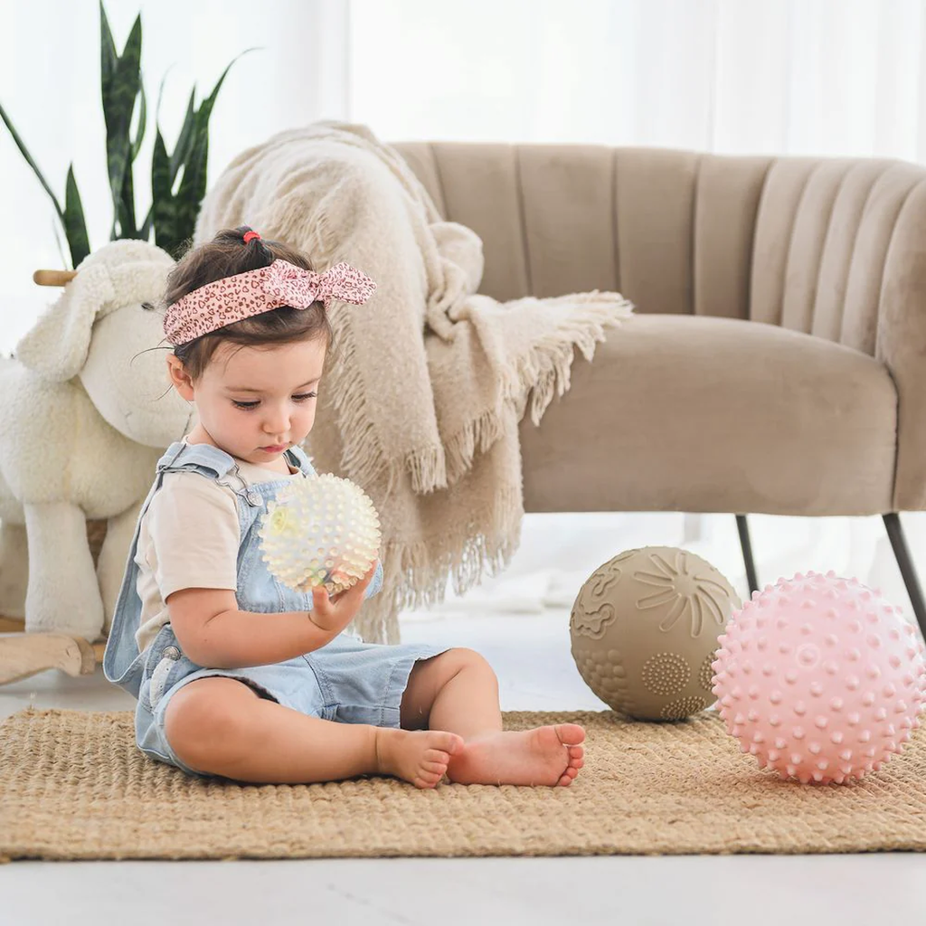 A toddler in denim overalls and a pink headband sits on a woven rug, holding a textured ball. Nearby are plush toys and soft furniture, creating a cozy scene.