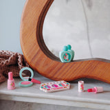 Wooden pretend makeup set with pink lipstick, eyeshadow palette, and teal perfume bottle by a curved mirror.