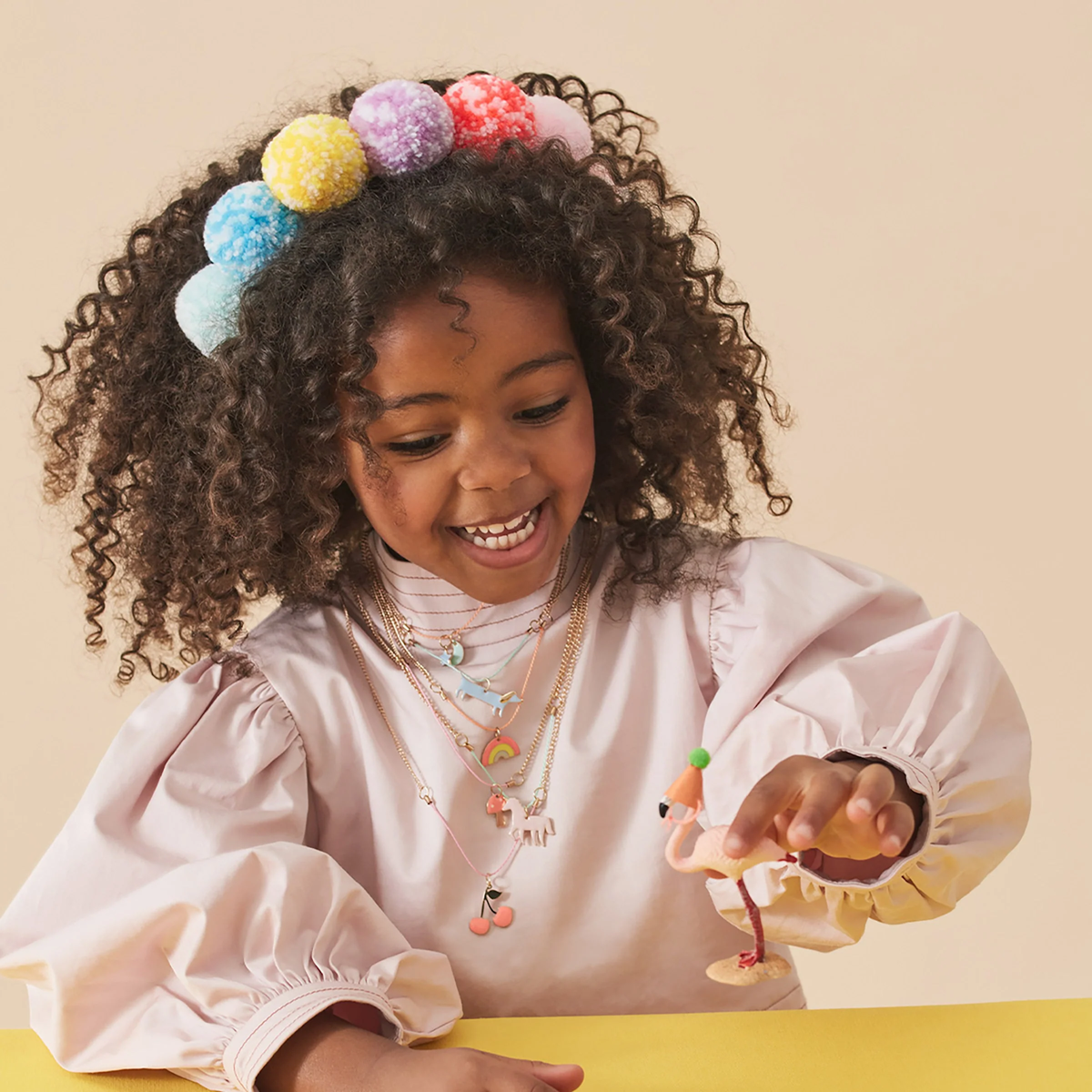 A young girl wearing a colorful pom-pom headband and multiple necklaces while playing with a flamingo toy.