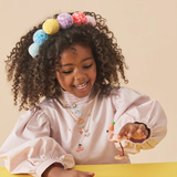A young girl wearing a colorful pom-pom headband and multiple necklaces while playing with a flamingo toy.