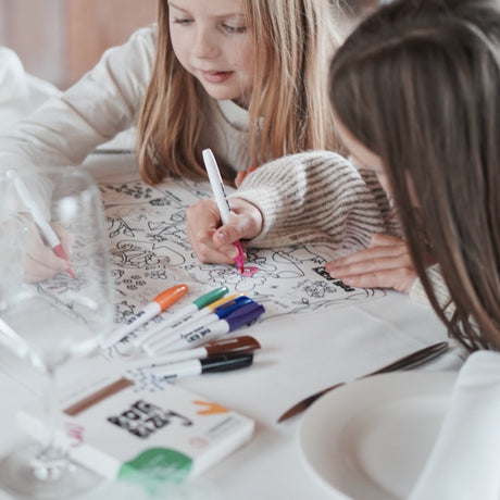 Two girls coloring a large activity mat with markers at a dining table.