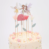 Frosted cake decorated with paper fairy toppers, tissue paper flowers, and a pink toadstool on wooden picks.