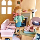 A wooden doll stands in a cozy dollhouse living room near a pink sofa and a tea set.