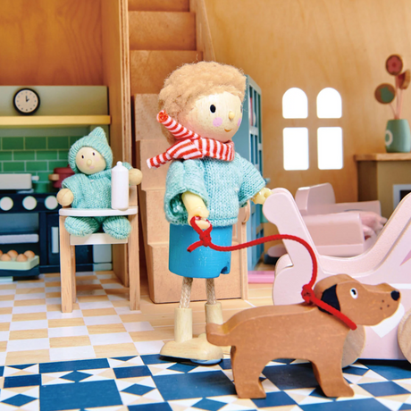 A wooden doll holding a leash walks a toy dog inside a dollhouse next to a baby in a high chair.