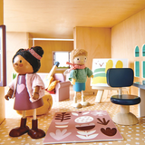 Two wooden dolls stand in a dollhouse room with an easel, office chair, and colorful rug.