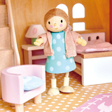 A wooden doll in a polka-dot dress and cardigan stands in a dollhouse bedroom near a bed and chair.