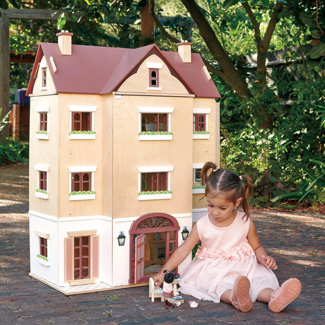 Young girl sitting outdoors playing with a doll next to the open front door of a wooden dollhouse.