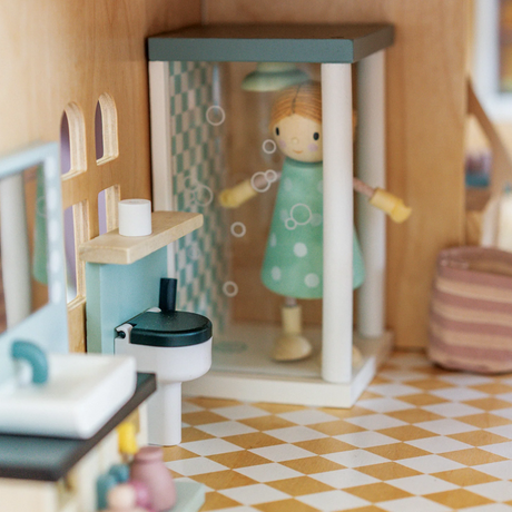 A wooden doll in a polka-dot dress stands inside a miniature dollhouse shower stall.