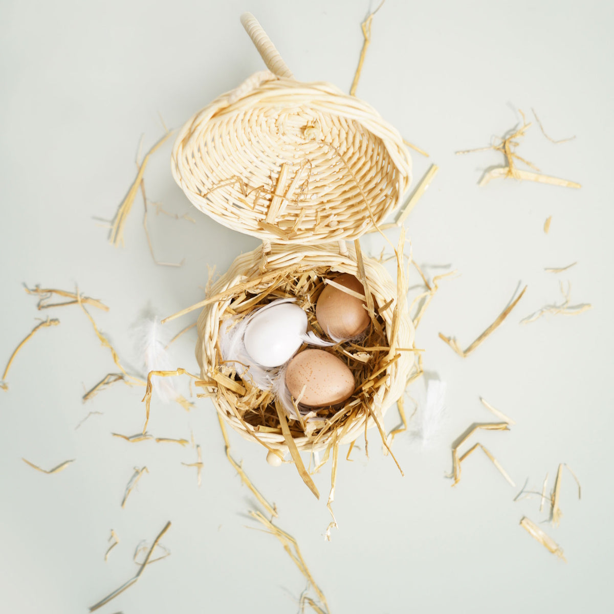 Top-down view of toy wooden eggs in a small woven basket with straw and feathers.