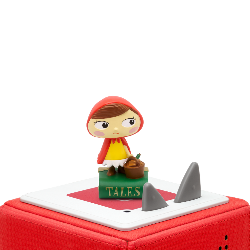 A cheerful Little Red Riding Hood figurine stands on a green book titled "Tales" audiobook in a white background. 