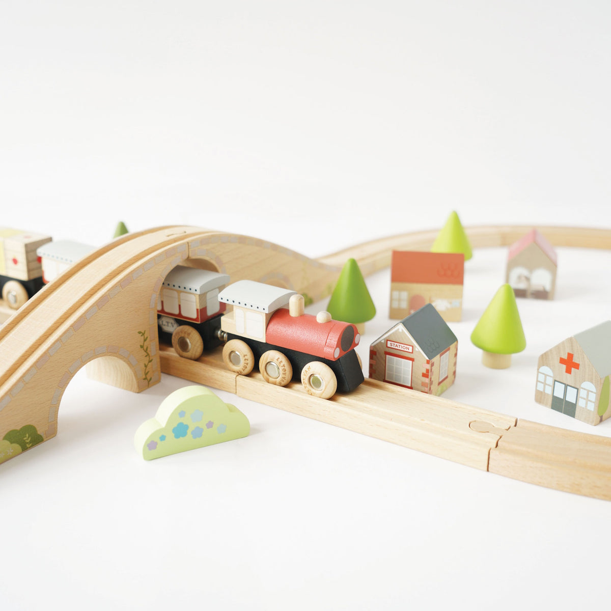 Wooden train passing under a bridge on a track set with toy buildings and trees.