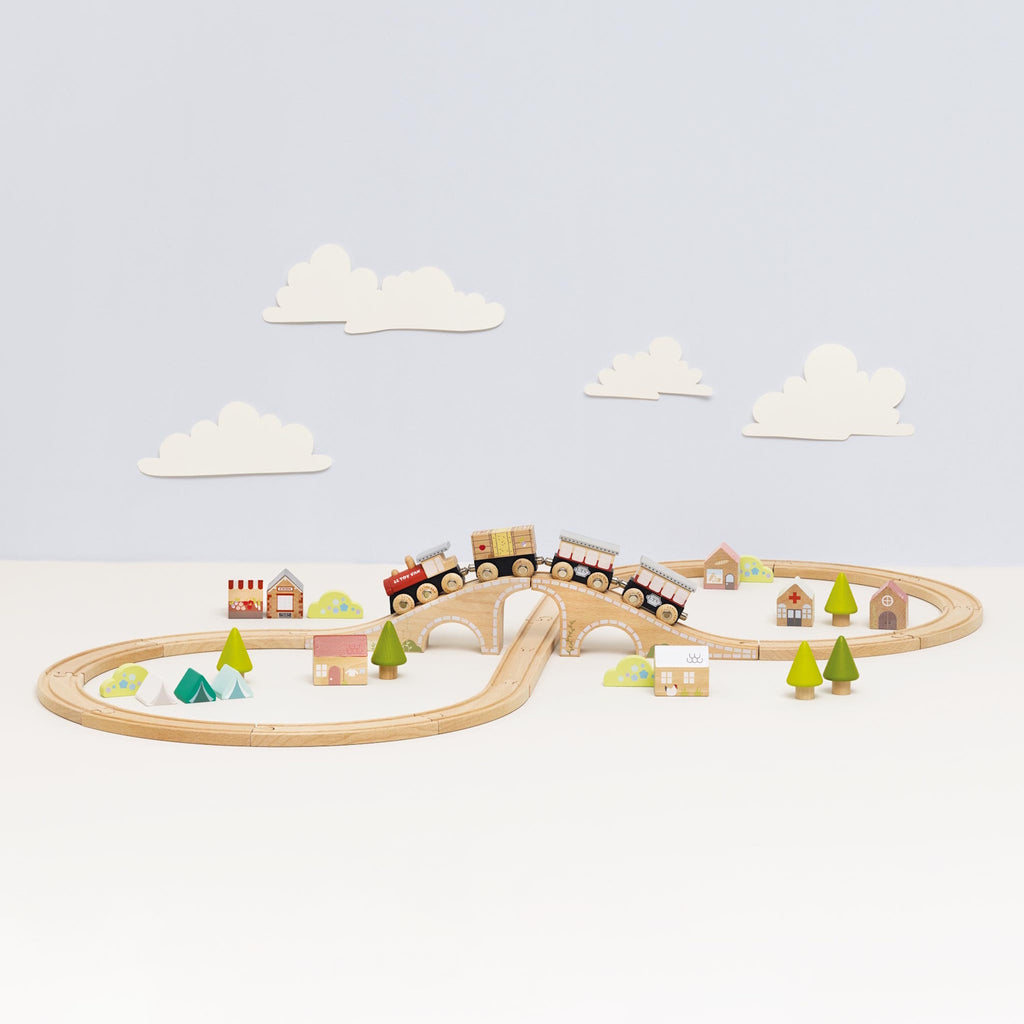 Wooden toy train set with a bridge and winding tracks is surrounded by small wooden trees and houses, creating a playful village scene.
