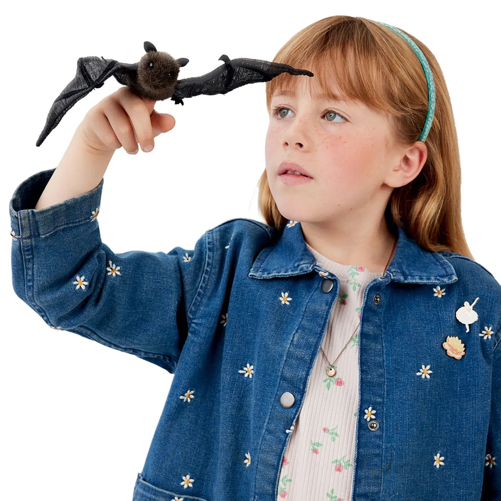 A young girl wearing a denim jacket with daisy patterns holds a small black bat on her finger. She looks at it with curiosity, creating a whimsical tone.