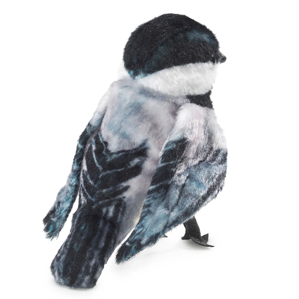 A plush bird toy with realistic black, white, and blue-gray feathers, facing away. It stands on two small, dark legs against a plain white background.