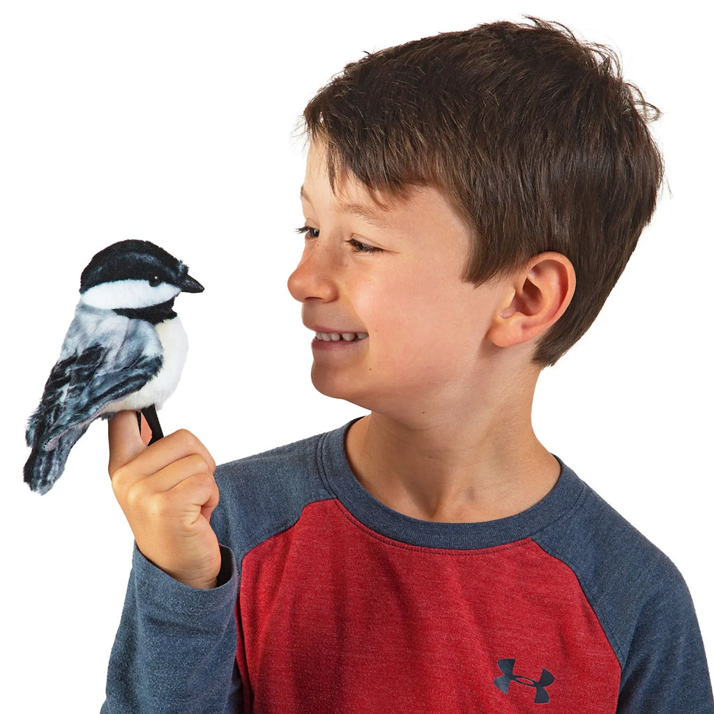 A young boy in a red and blue long-sleeve shirt holds a plush bird on his finger. He smiles warmly at the bird against a plain white background.