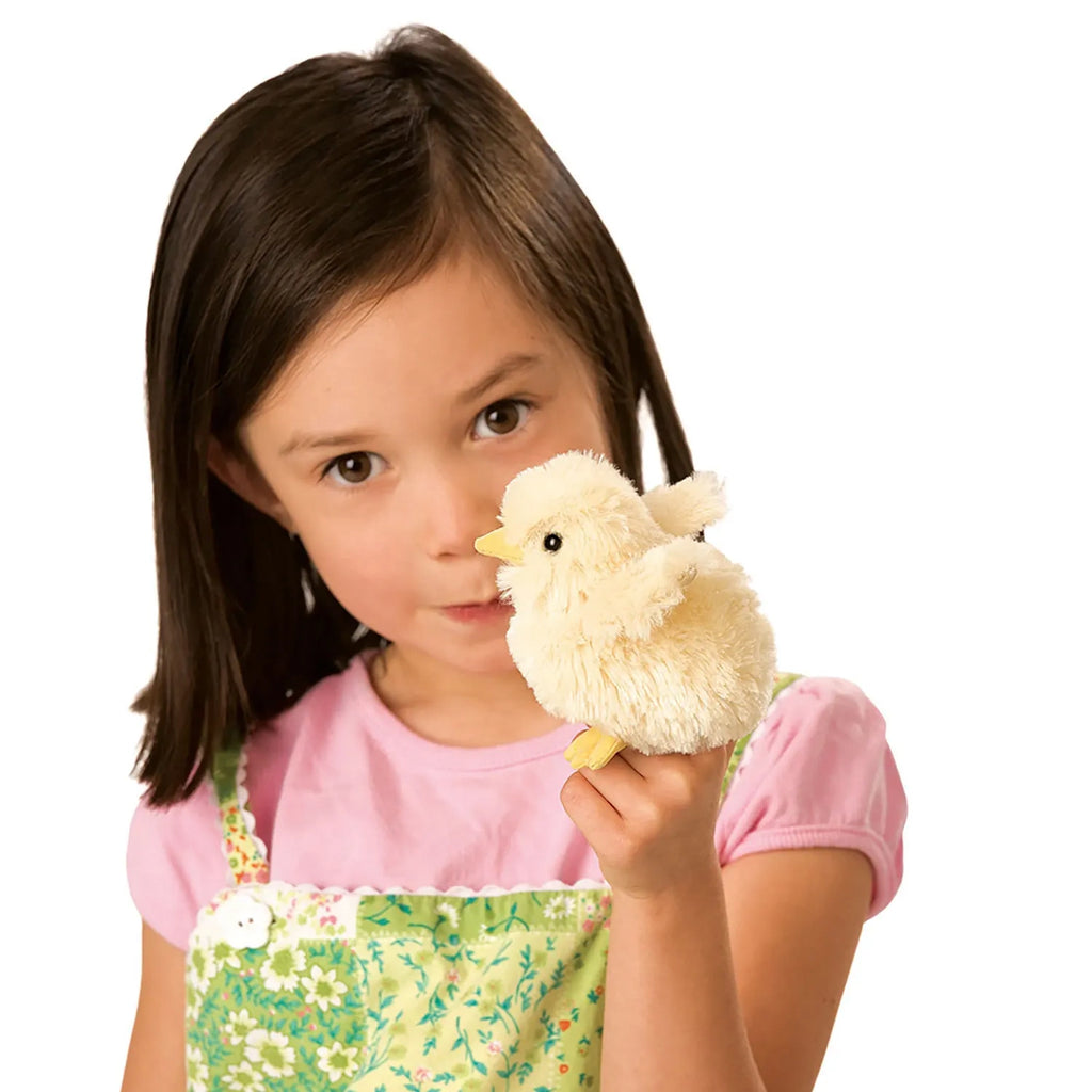 A young girl in a pink shirt holds a fluffy yellow chick toy up to her face, with a curious expression. She wears a green floral dress.