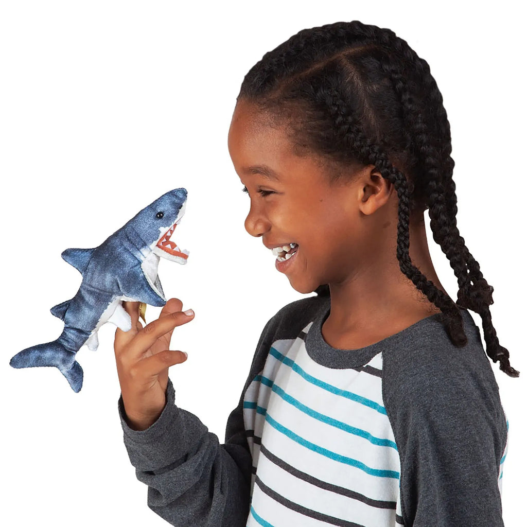 A smiling child with braided hair plays with a shark hand puppet, wearing a striped shirt. The scene is joyful and playful.