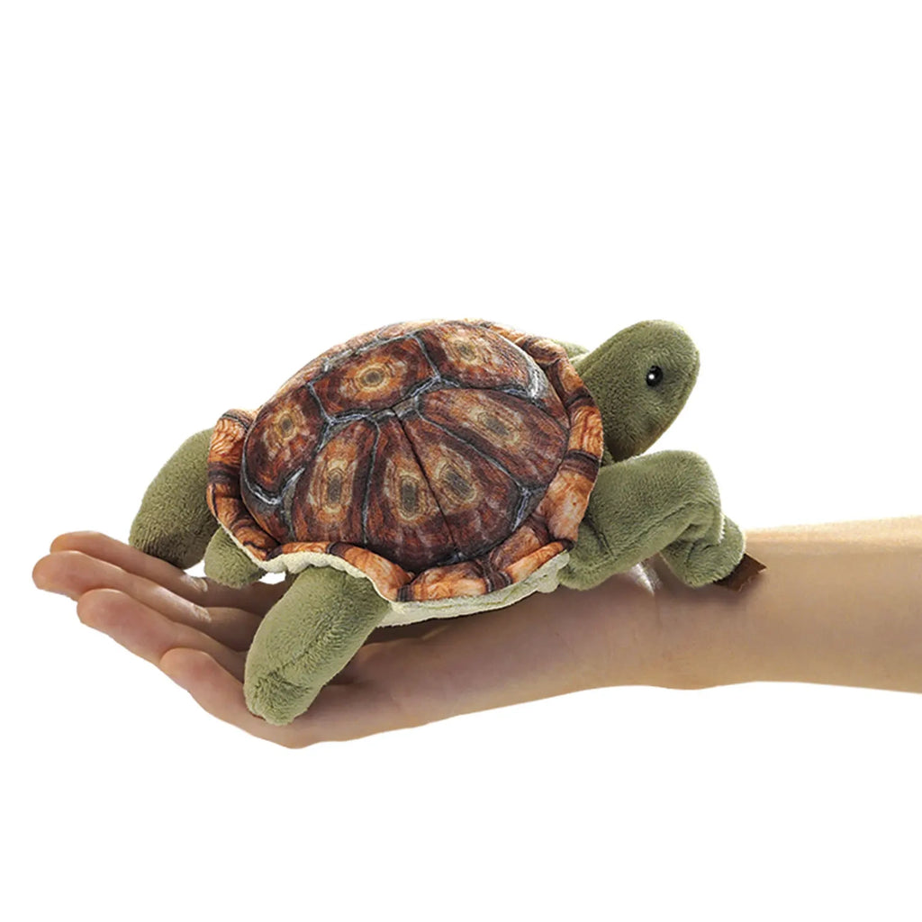 A plush turtle with a detailed brown shell rests on a person's open palm. The turtle's soft, green fabric and cute expression evoke warmth and playfulness.