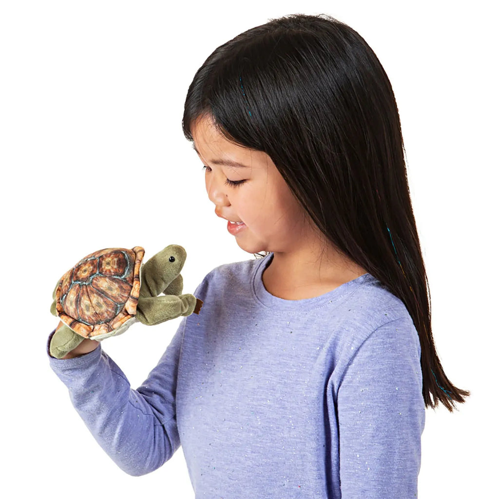 A young girl with long hair smiles while holding a turtle puppet on her hand. She wears a light purple shirt. The scene is joyful and playful.