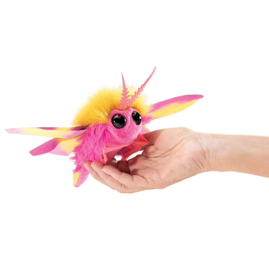 A small, colorful plush toy resembling a pink and yellow moth with large eyes is held gently in a person's open hand, conveying a playful tone.