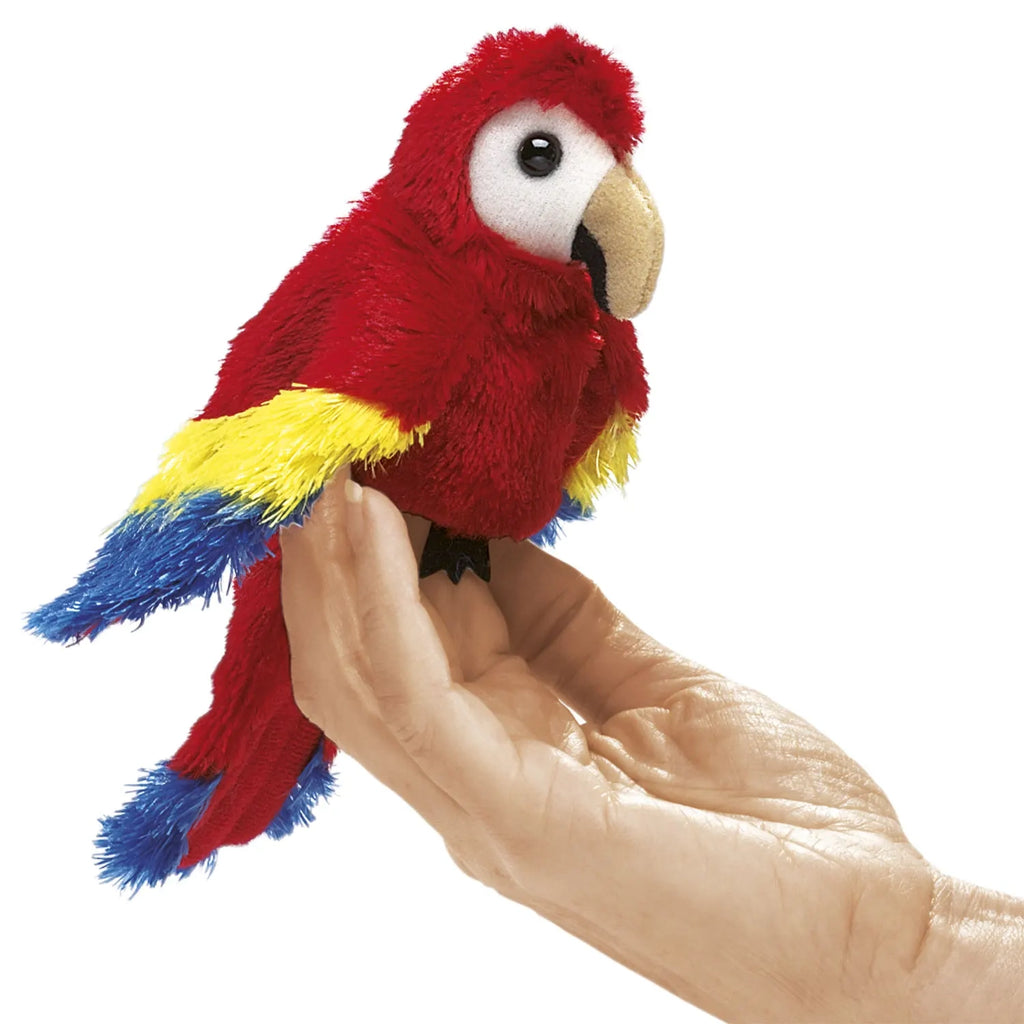 A plush parrot toy in vivid red, blue, and yellow sits on a person's hand against a white background, conveying a playful, whimsical tone.