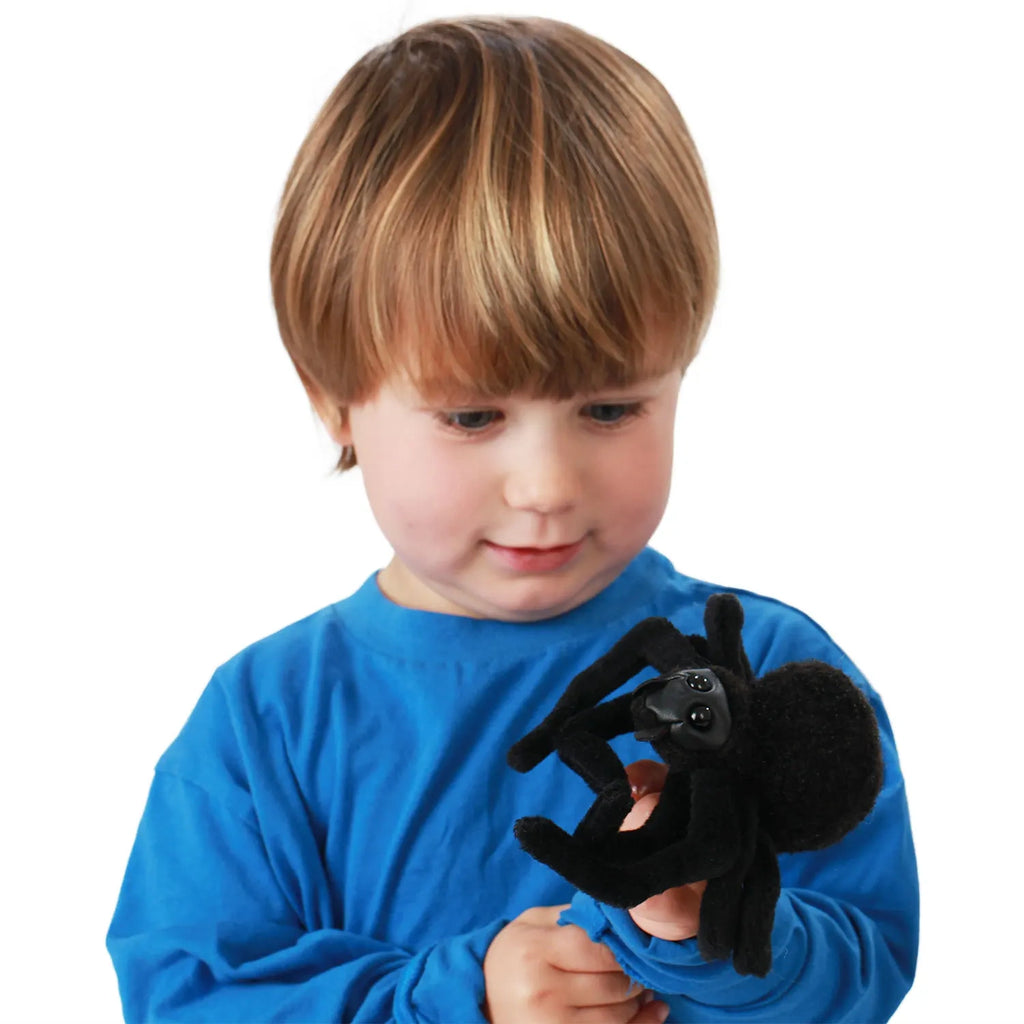 A young child in a blue shirt holds a fuzzy black spider toy in their hand, looking at it with curiosity. The background is plain white.