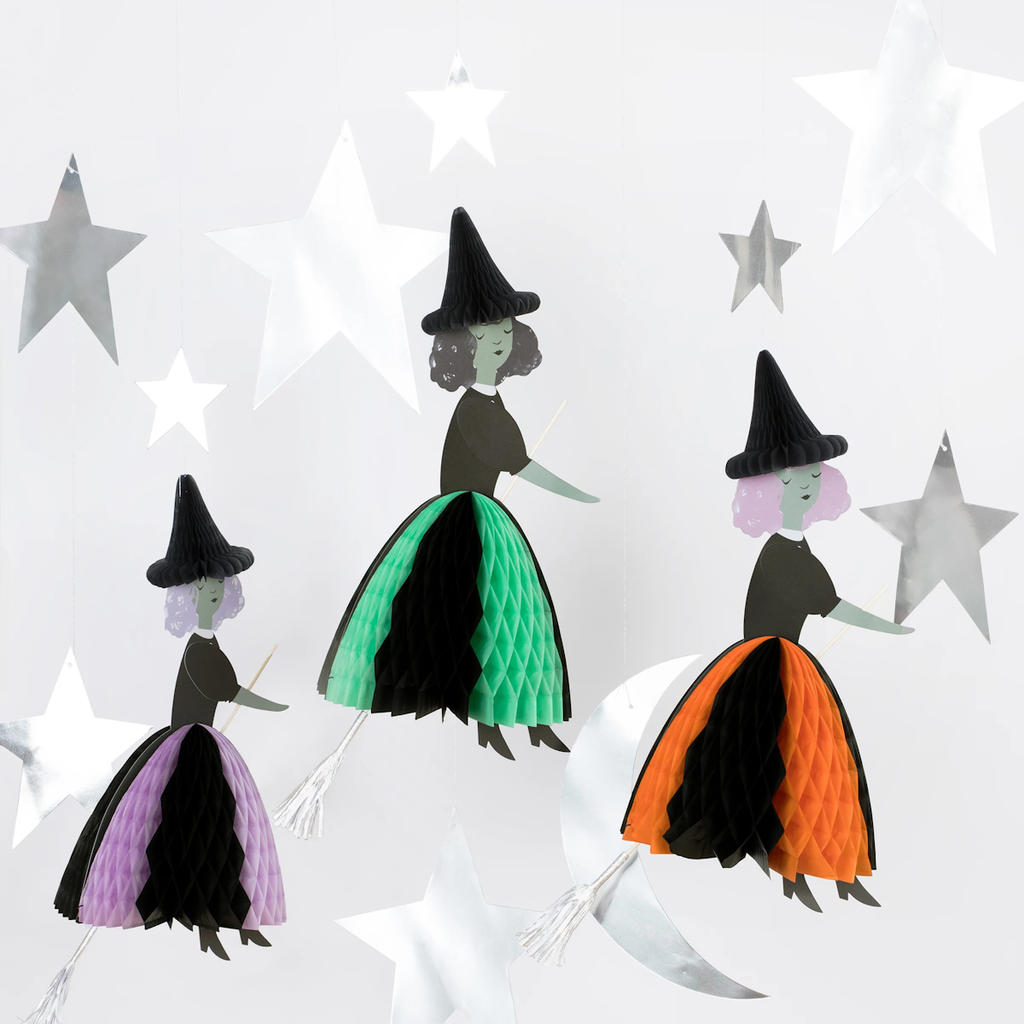 Three hanging witch decorations with paper honeycomb skirts in orange, green, and purple. They have black hats and brooms, surrounded by silver stars.