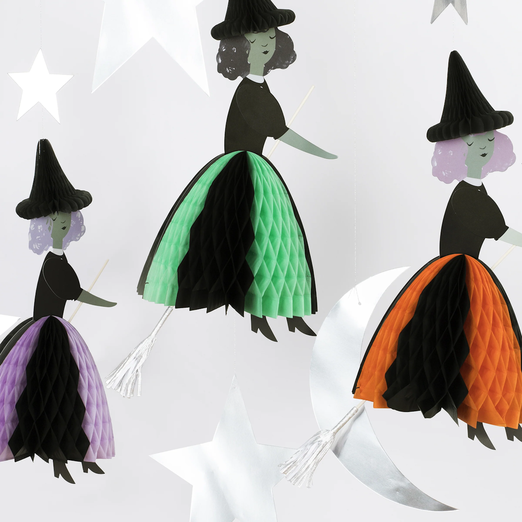 Three paper witches in colorful skirts and black hats hang among white stars and a crescent moon. The scene is whimsical and festive.