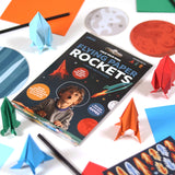 Flying Paper Rockets activity book kit surrounded by folded paper rockets, stickers, and planet cutouts.
