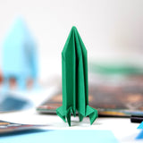 Green origami rocket standing upright on a white surface with craft supplies in the background.