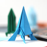 A blue folded origami rocket standing upright with a green paper rocket blurred in the background.