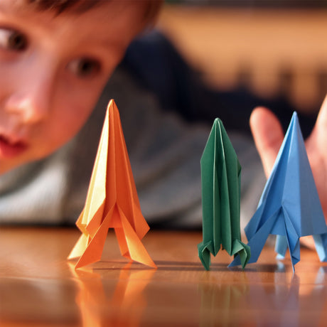 Young boy looking closely at three colorful origami rockets standing upright on a wooden table.