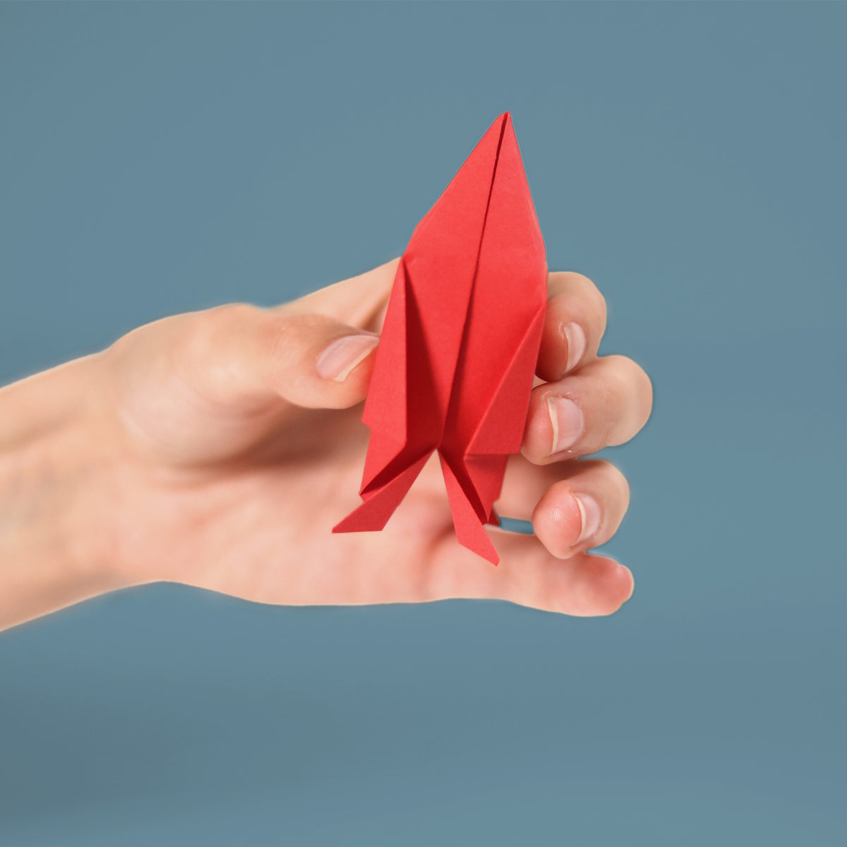 A hand holding a folded red paper rocket against a plain blue background.