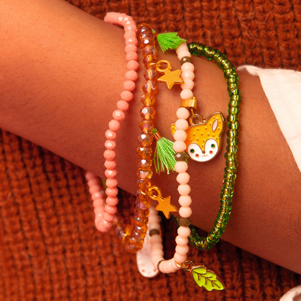 Five colorful beaded bracelets adorn a wrist, featuring charms of a fox, stars, and a leaf, set against a cozy brown knitted fabric. Playful and vibrant.