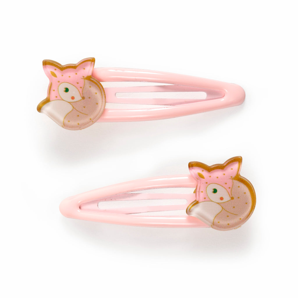 Two pink hair clips shaped like foxes with polka dots and green eyes, laid on a white background. The tone is playful and whimsical.
