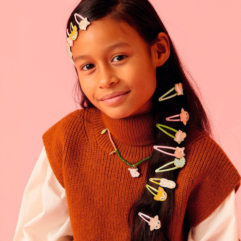 A young girl with long dark hair, wearing a rust-colored sweater and white shirt, smiles softly. Her hair is adorned with colorful cartoon hair clips, matching a whimsical necklace. The soft pink background adds a playful tone.