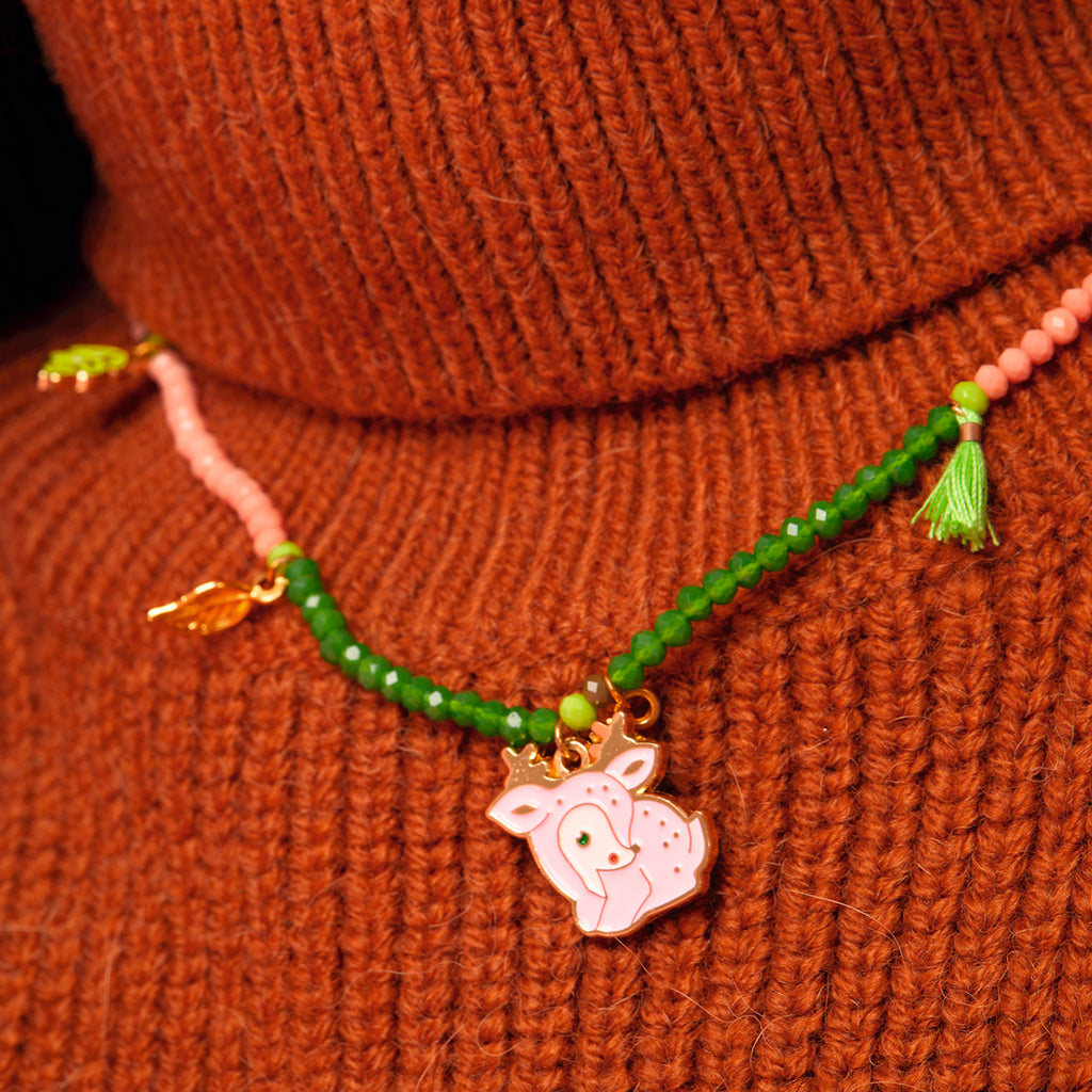 A colorful necklace with a pink deer charm and green beads hangs around a neck wearing an orange knit sweater, conveying a playful and cozy vibe.