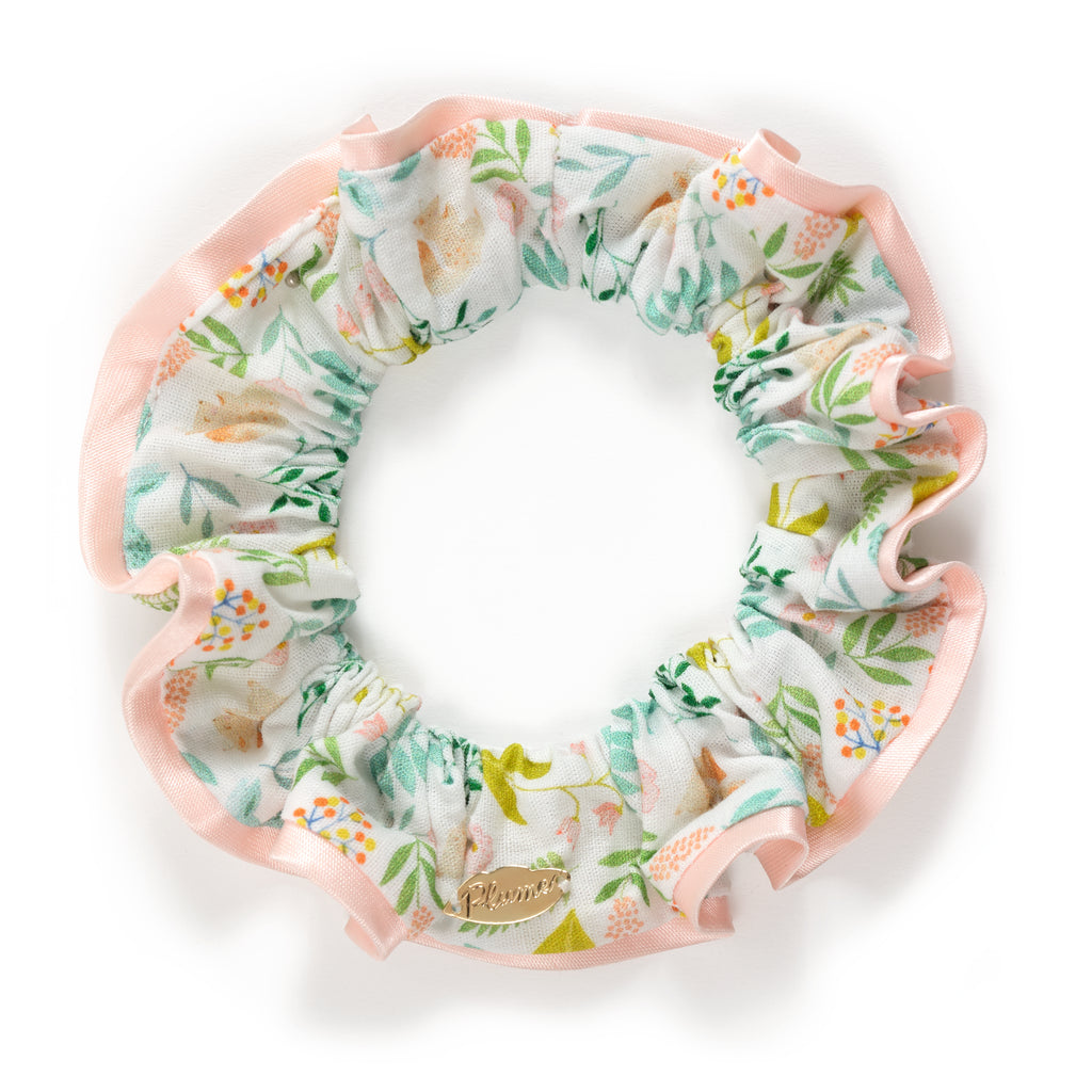 Floral scrunchie with pink ruffled edges, featuring green leaves and orange flowers. It includes a small, gold metal tag on a white background.