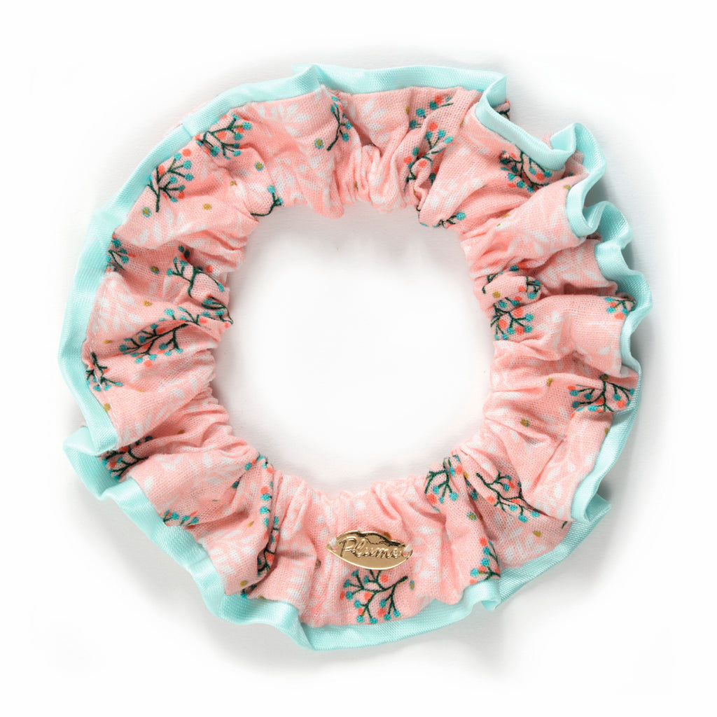 A pink fabric scrunchie with blue floral patterns and a turquoise border. It has a gold emblem attached, conveying a soft, feminine look.